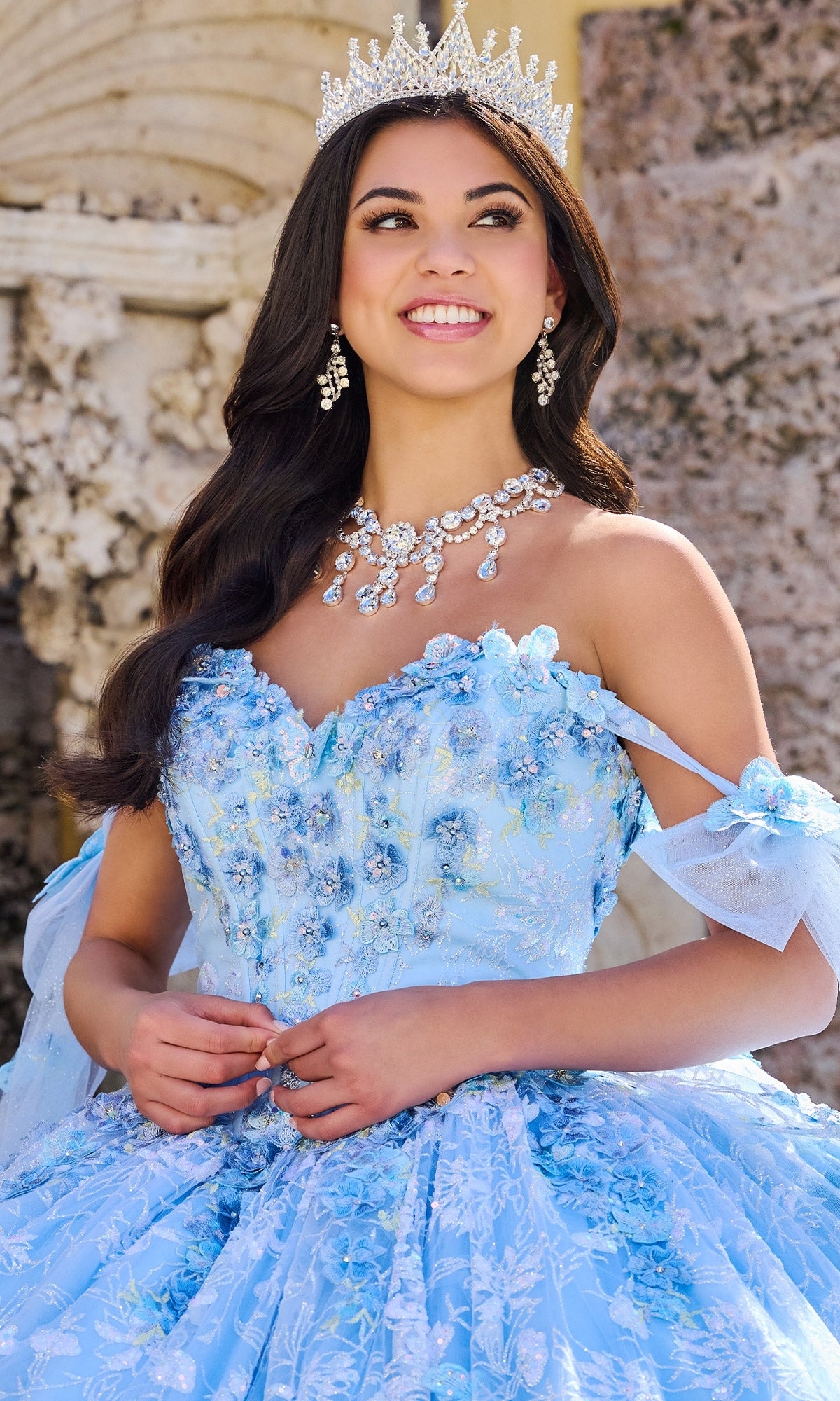 A young woman smiles outdoors, wearing the Princesa PR30135 3D Floral Quinceanera Dress with a corset bodice, sparkling silver tiara, and statement jewelry.